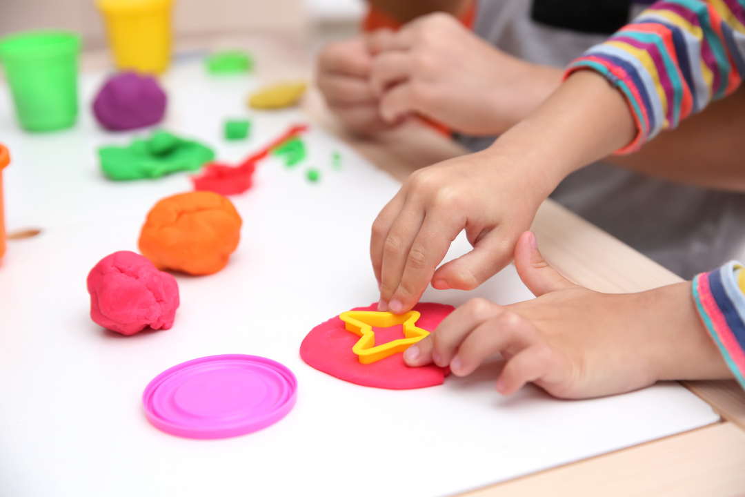 Small child hands are seen placing a yellow star shape cutter into a circle of red playdough.
