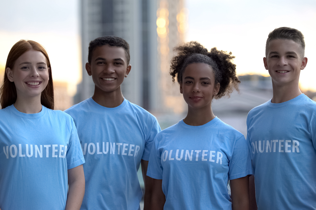 Four teenagers stand in a row facing the camera. They are smiling and wearing matching blue shirts that say volunteer.