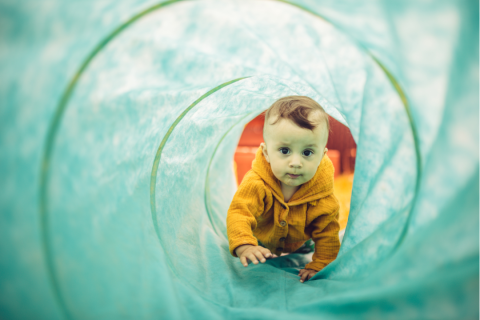 A small boy in a yellow shirt crawls through a blue green tunnel.
