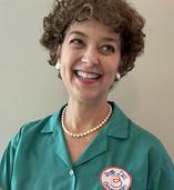 Image is a photo of a white woman with short curly brown hair wearing a green button up shirt with a pearl necklace and earrings. The photo is of historian Leslie Goddard appearing as Julia Child.