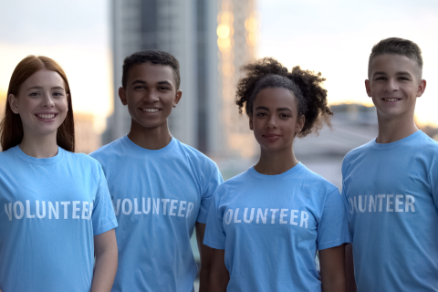 Four teenagers stand in a row facing the camera. They are smiling and wearing matching blue shirts that say volunteer.