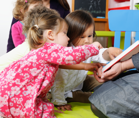 A picture of young toddlers looking at pointing at an open picture book that is being held by an adult.