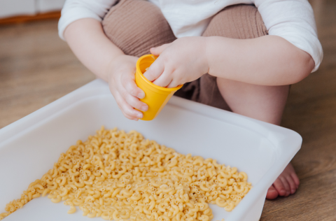 A child's arms and legs are seen next to a white plastic bin with dried macaroni noodles inside. The child is scooping the noodles into a small yellow cup.