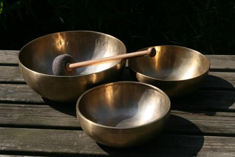 Three metal bowls on a wooden table with a mallet that looks like a dowel with a small knob on one end and a burnt marshmallow on the other, which is resting diagonally in one of the singing bowls.