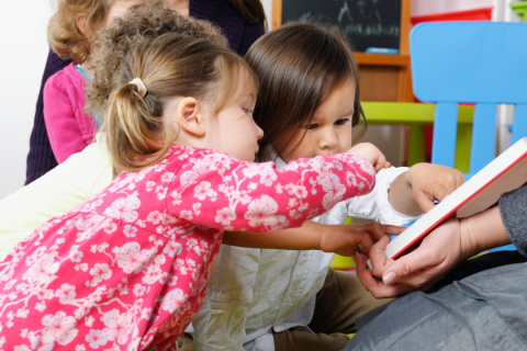 A picture of young toddlers looking at pointing at an open picture book that is being held by an adult.