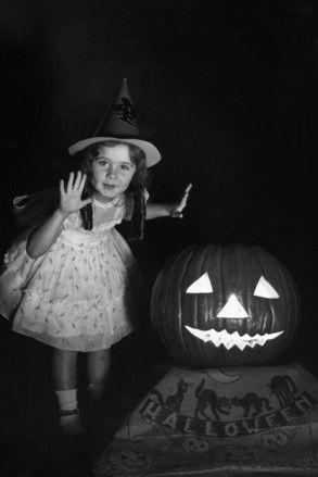 Black and White photo of a young girl with dark hair in pigtails and a light colored dress, standing next to a lit jack-o-lantern.