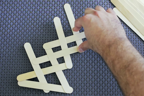 A hand adjusts some plain colored popsicle sticks to create a stick bomb