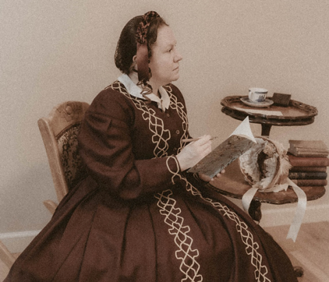 Photo is a sepia toned image of historical reenactor Laura Keyes portraying Elizabeth. She is sitting in a chair and dressed in a dark colored dress from the 1860s.