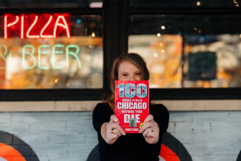 photo of Molly Page in front of a pizza parlor, holding a copy of her book "100 Things to Do in Chicago Before You Die."