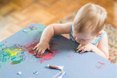 A small child is pushing around red paint with their hand on a large piece of blue paper and is already covered in paint.