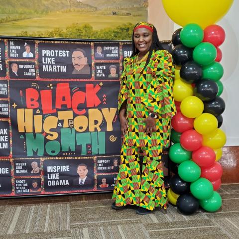 Image is of storyteller and educator Elizabeth Kahura, an African American woman dressed in traditional African apparel, standing in front of a Black History Month display. There is a column of balloons next to her in stripes of red, green, black, and yellow. 