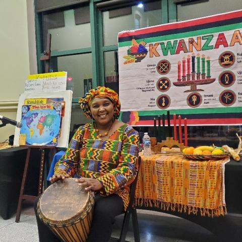 Shows storyteller and performer Elizabeth Kahura playing a drum in traditional african apparel. She is sitting in front of a Kwanzaa themed bulletin board display.