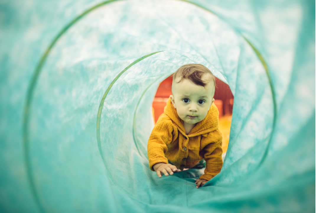 A small boy in a yellow shirt crawls through a blue green tunnel.