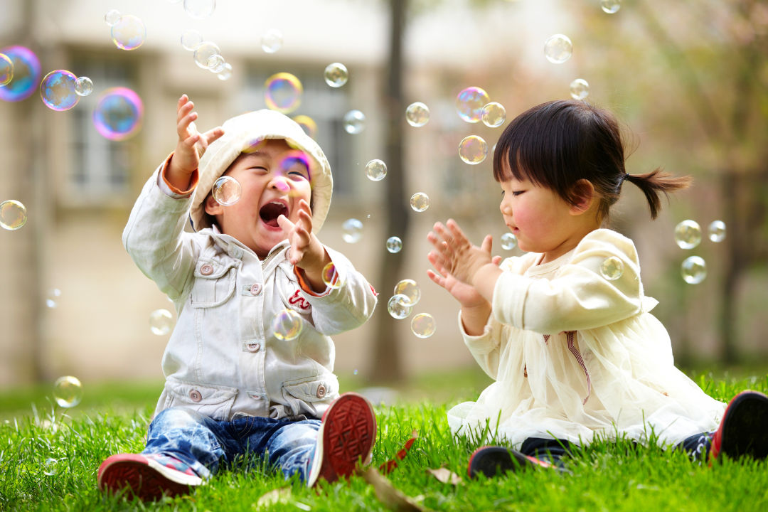 Two toddlers sit in the green grass playing with bubbles that are floating by.